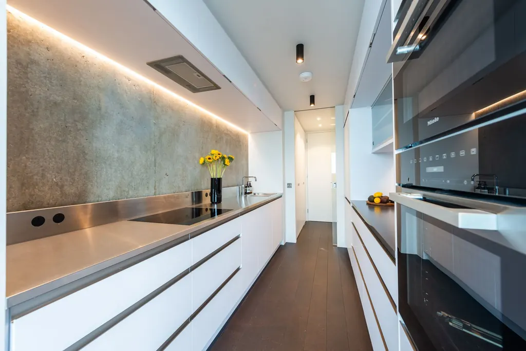 A long galley kitchen with white handleless units, stainless worktop and a concrete splashback lit from above